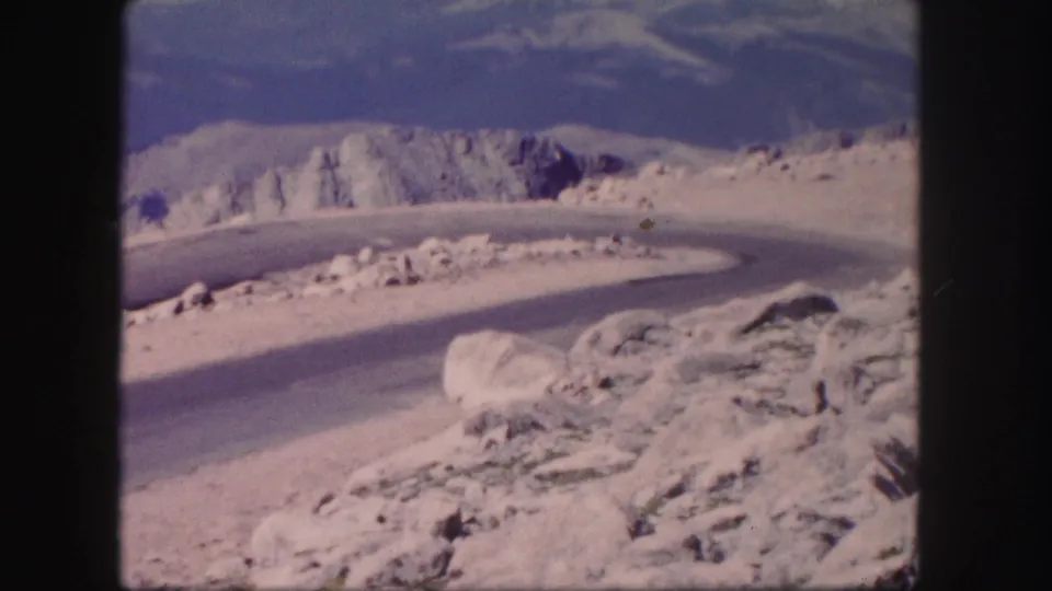 1958 Colorado 8mm frame of rocky roadside terrain, a winding mountain road, sparse vegetation, and distant ridges.
