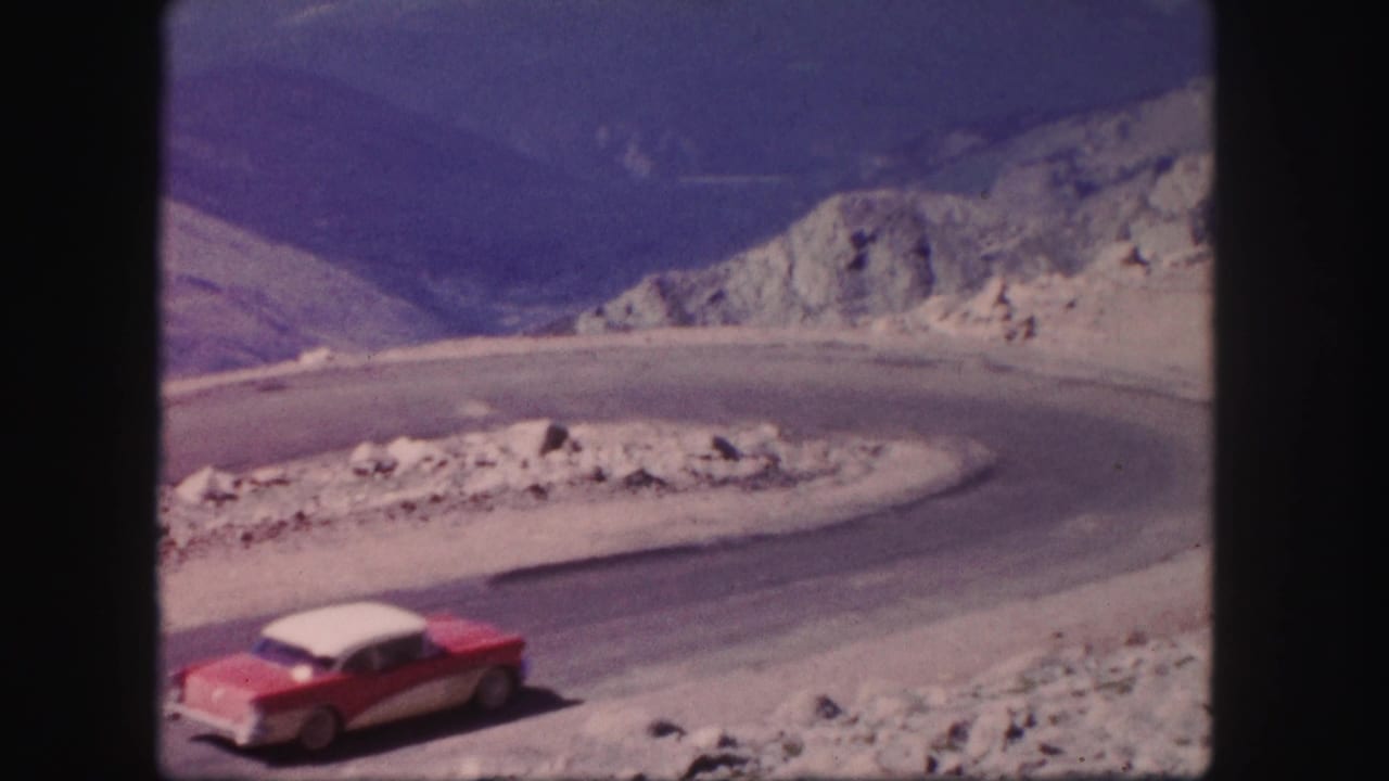 Red and white sedan entering a sharp mountain road curve below rocky slopes.