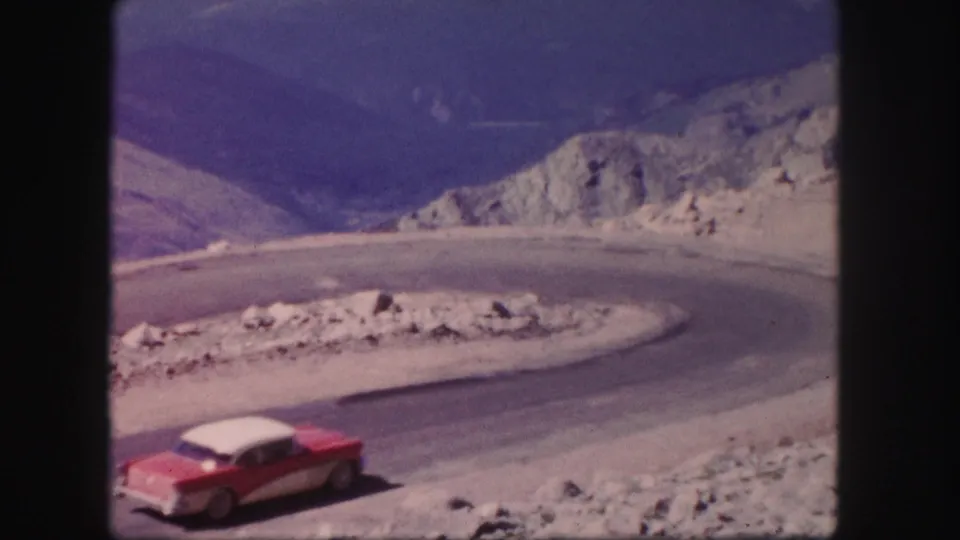 1958 Colorado home-movie frame of a red and white sedan rounding a hairpin road in a rocky mountain landscape.