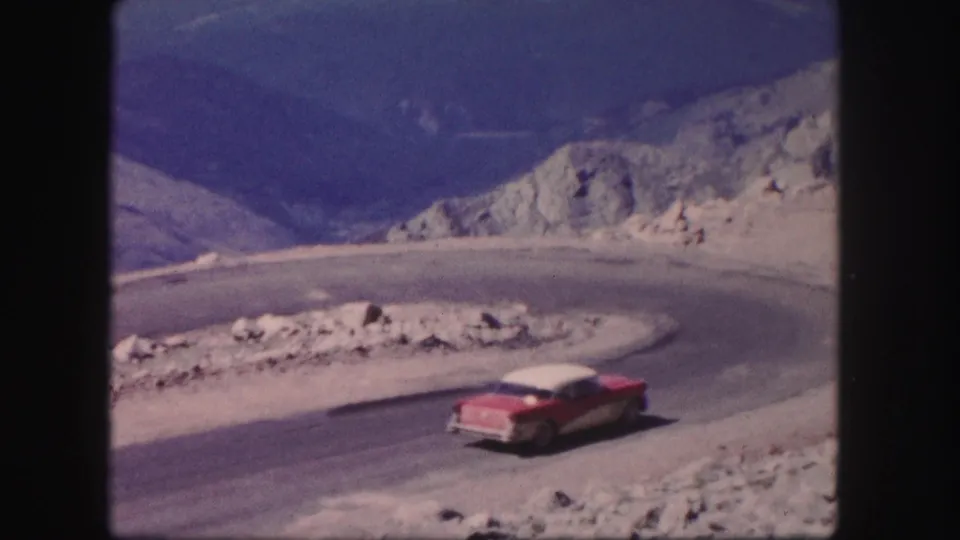 1958 Colorado 8mm frame showing a red and white sedan continuing along a curved mountain road with rocky terrain behind it.