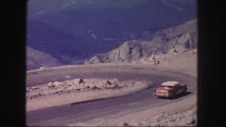 1958 Colorado home-movie frame of the sedan moving away around a mountain road curve amid pale rocks and distant slopes.