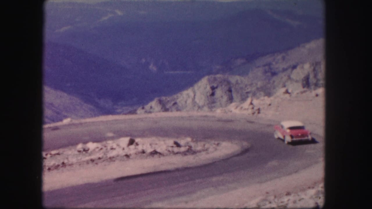 Red car continues around a paved mountain curve with distant slopes behind it.