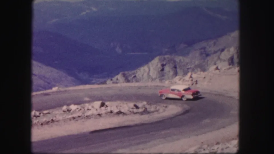 A mid-century red and white car is seen broadside while turning through the Colorado mountain road bend.