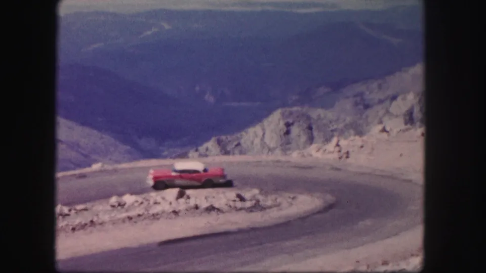 The red and white car continues out of the winding Colorado mountain road curve in grainy 8mm footage.