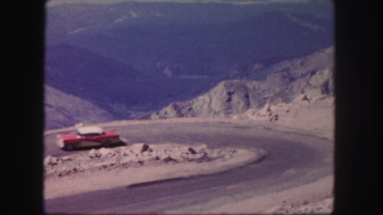 A red vintage car rounds a tight curve on a rocky Colorado mountain road in metadata-supported 1958 8mm footage.