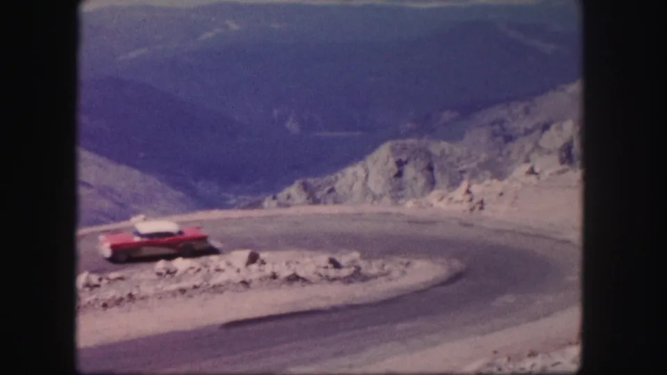 A red vintage car rounds a tight curve on a rocky Colorado mountain road in metadata-supported 1958 8mm footage.
