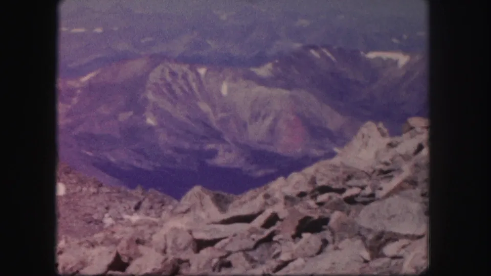 A wide mountain landscape in Colorado shows rocks in the foreground and layered ridges beyond, recorded on 8mm film in 1958.