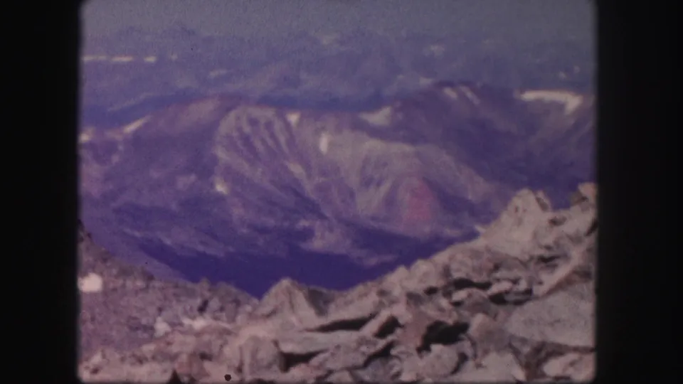 A rocky Colorado overlook faces distant ridges and a hazy valley in a soft, color-shifted 1958 8mm home-movie frame.