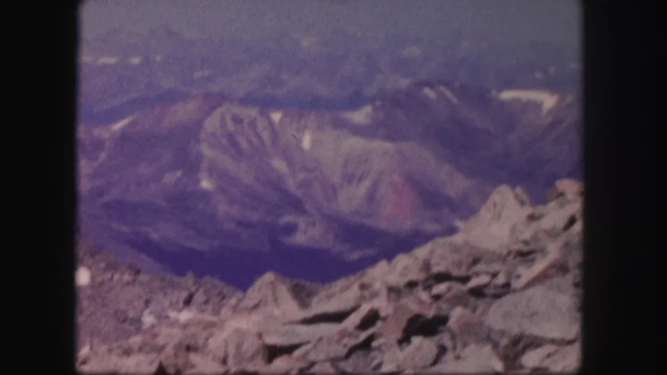 1958 Colorado 8mm frame with visible grain, gray rocks in front, and blue-purple mountain ridges in the distance.