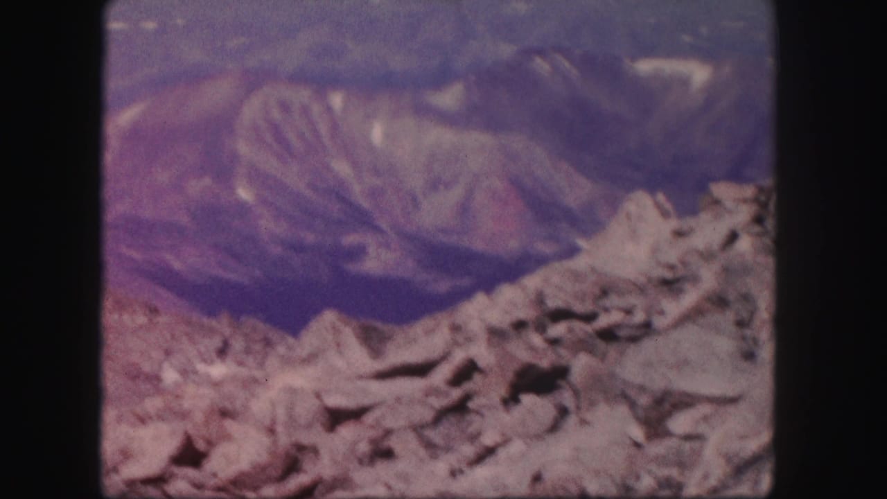 1958 Colorado home-movie frame with jagged foreground rocks, distant ridgelines, and strong purple-blue film fading.