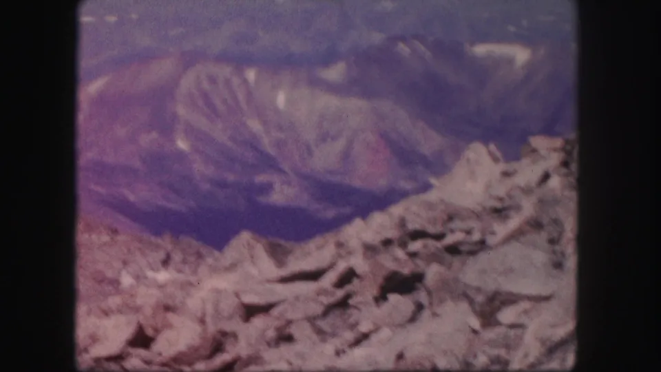 1958 Colorado home-movie frame with jagged foreground rocks, distant ridgelines, and strong purple-blue film fading.