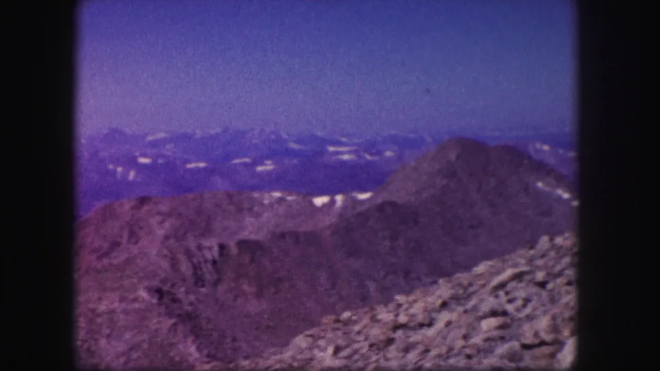 1958 Colorado 8mm view across a rocky alpine slope toward rounded ridges and far snow-streaked mountain peaks.