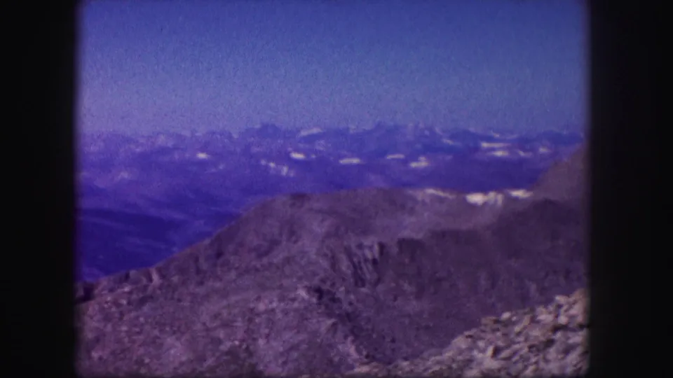 1958 Colorado 8mm frame showing a rocky high ridge, layered mountain ranges, and scattered snow patches beneath a blue sky.