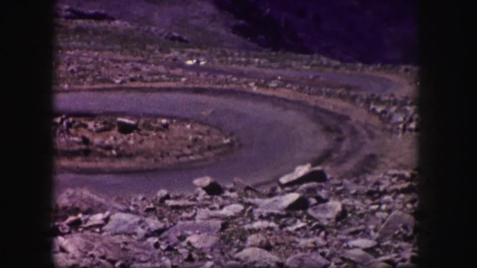 1958 Colorado 8mm frame looking over rocks toward a sweeping paved road curve in sparse desert terrain.