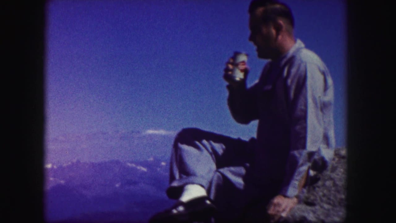 Man seated on a rocky mountain edge drinking from a can against a blue sky.
