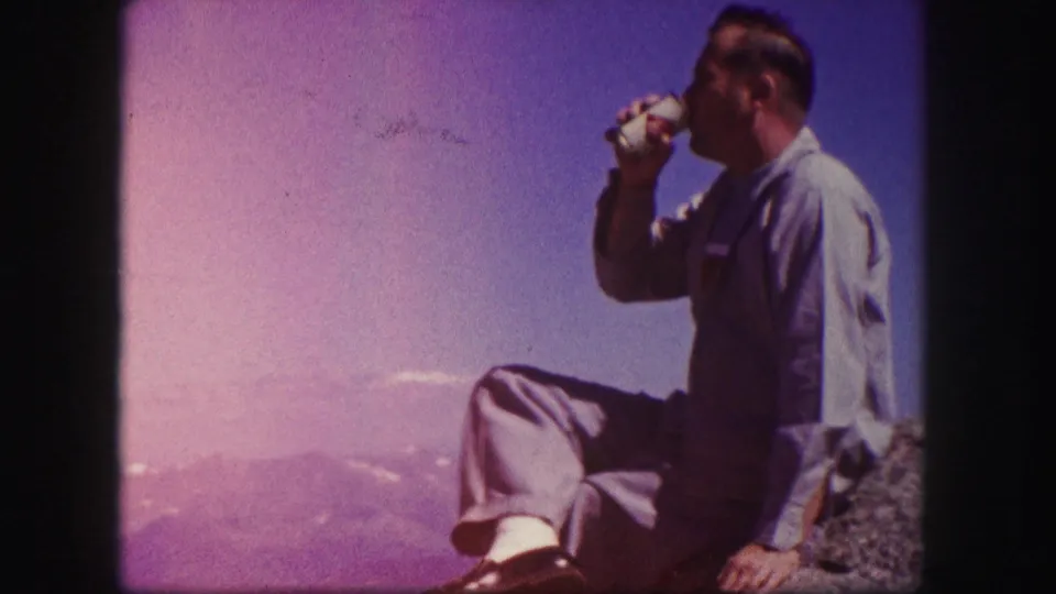 1958 Colorado 8mm frame showing a side view of an adult man drinking from a can while seated on a rocky overlook above distant mountains.