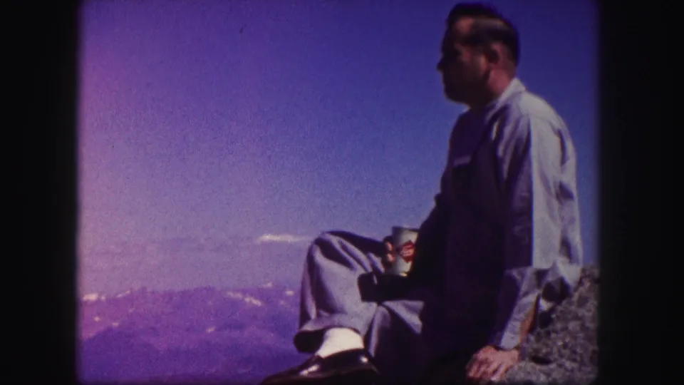 1958 Colorado 8mm frame of an adult man holding a can while seated at a rocky mountain overlook with distant ridges under blue sky.