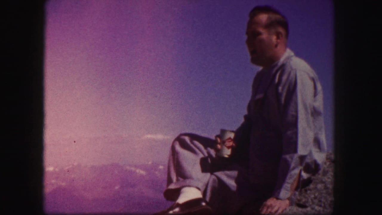 Man in blue shirt sits on a rock holding a beverage can with mountains behind him.