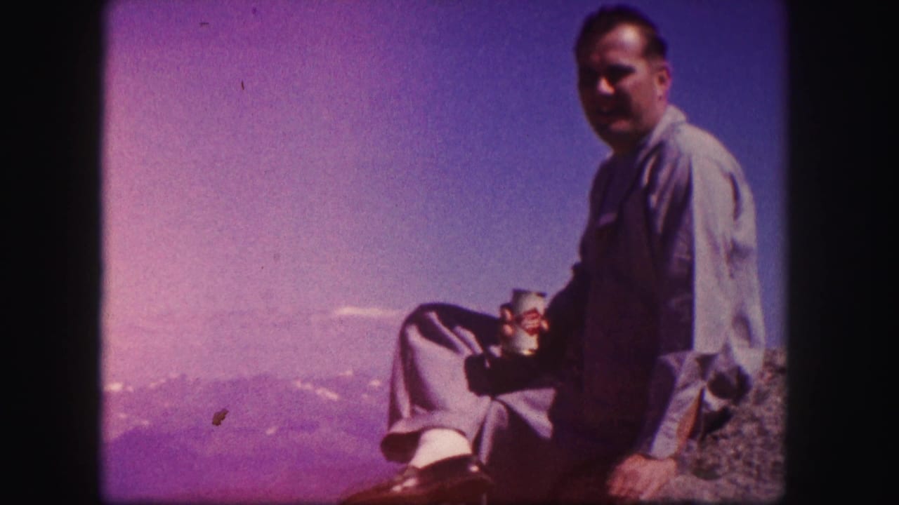 A man seated on a Colorado rocky overlook turns toward the camera, holding a beverage can beneath a clear blue sky.