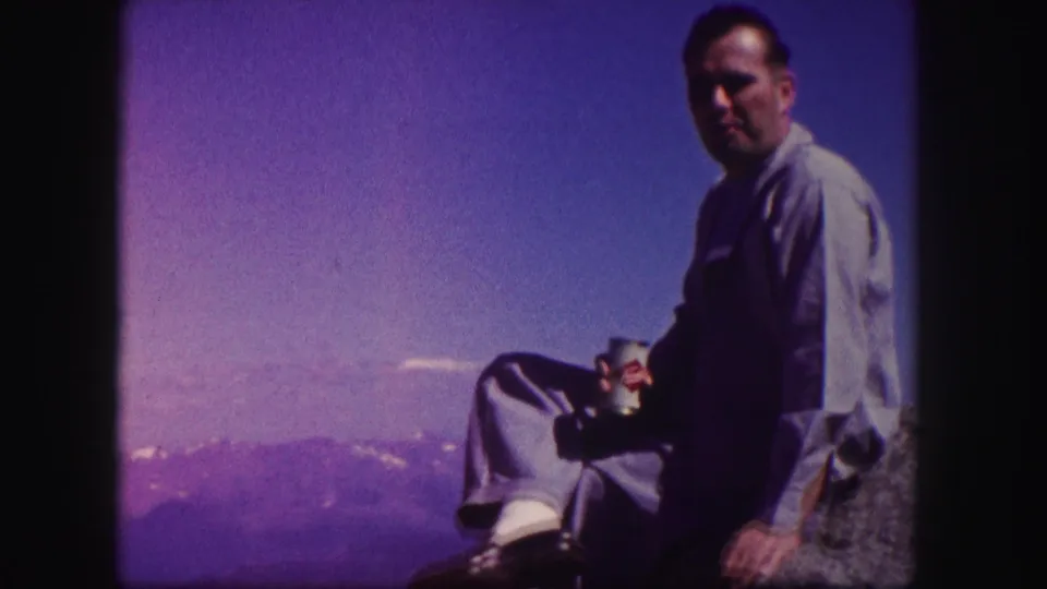The 1958 home-movie frame shows a seated man in a blue shirt holding a can, with Colorado mountain ridges visible behind him.