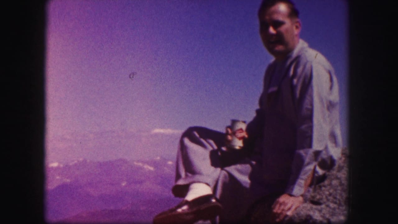 Man seated on a rocky cliff with mountains and sky behind him.