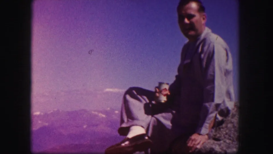 Adult man in Colorado, 1958, seated on rock with a cup, distant mountains, and purple-blue sky behind him.