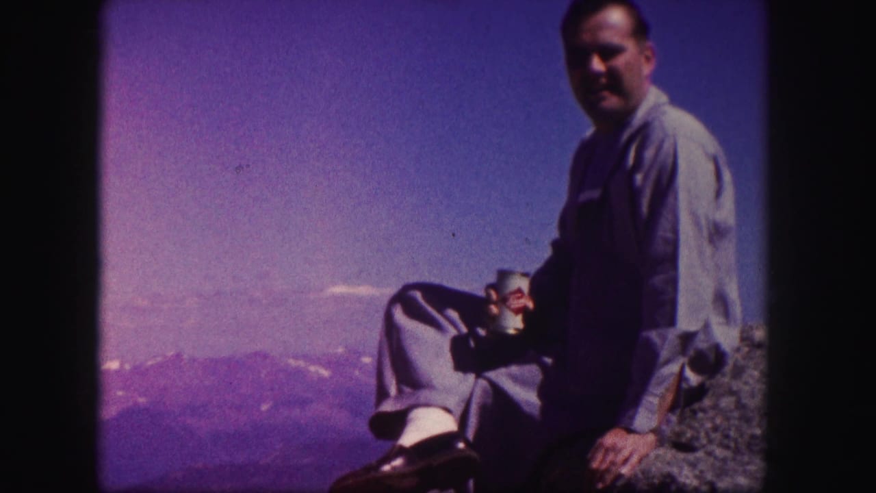 Seated man holding a cup on a rocky overlook above distant mountains.