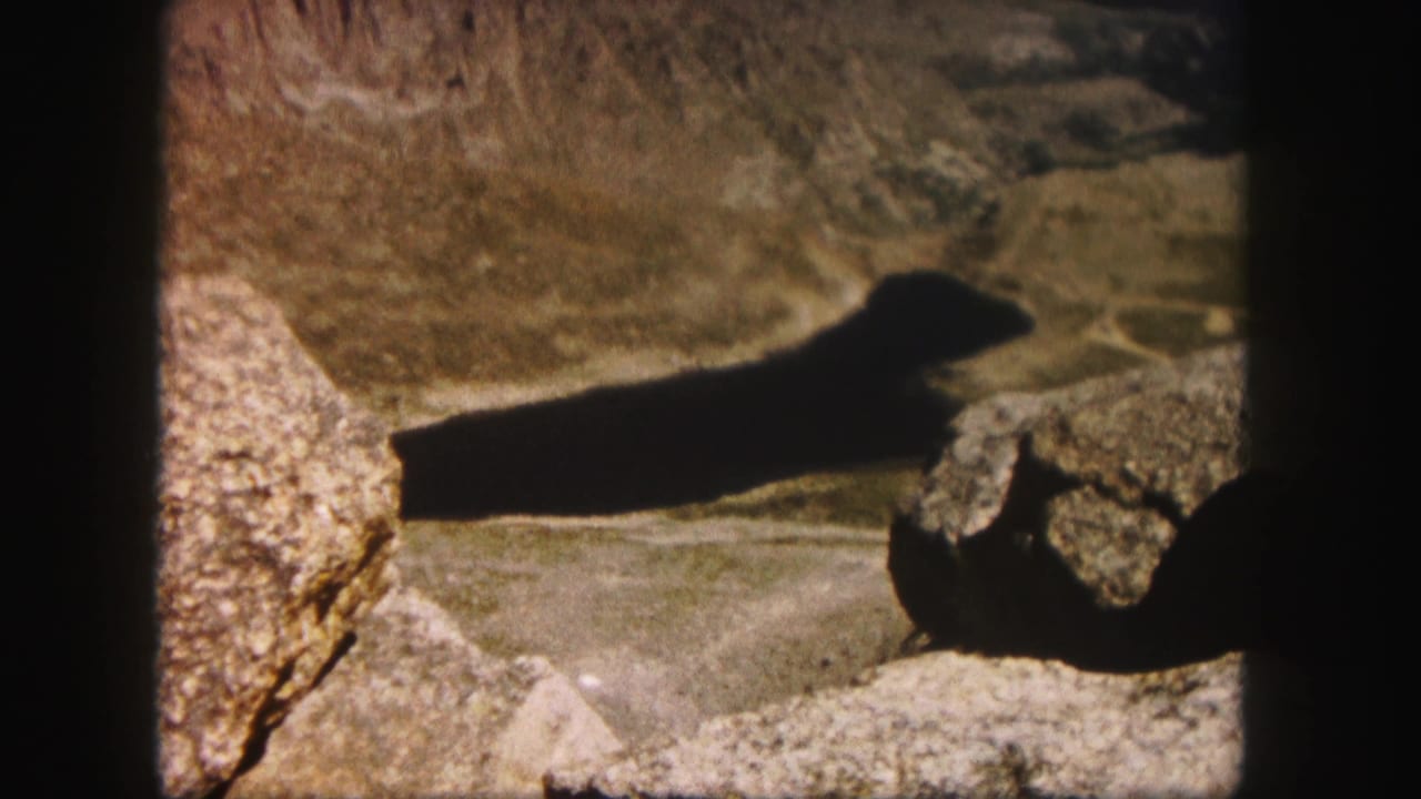 1958 Colorado 8mm frame showing a dark elongated lake or reservoir below rocky foreground boulders and arid mountain slopes.