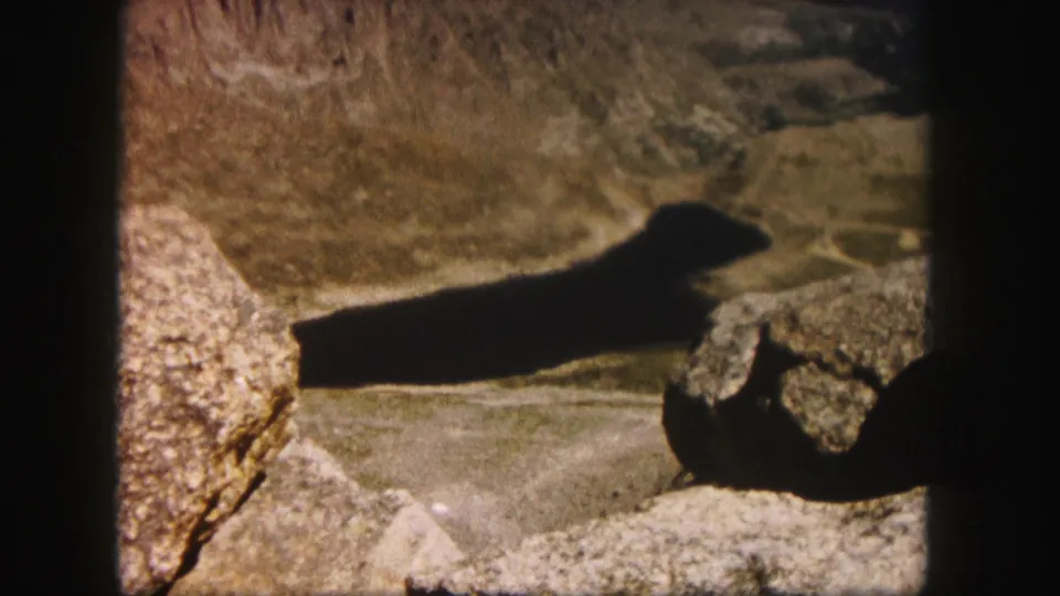 1958 Colorado 8mm frame showing a dark elongated lake or reservoir below rocky foreground boulders and arid mountain slopes.