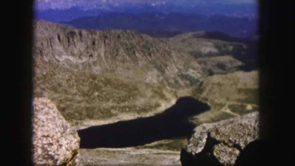 1958 Colorado home-movie still of a dark lake or reservoir between rocky slopes, with boulders in the foreground and distant hills beyond.