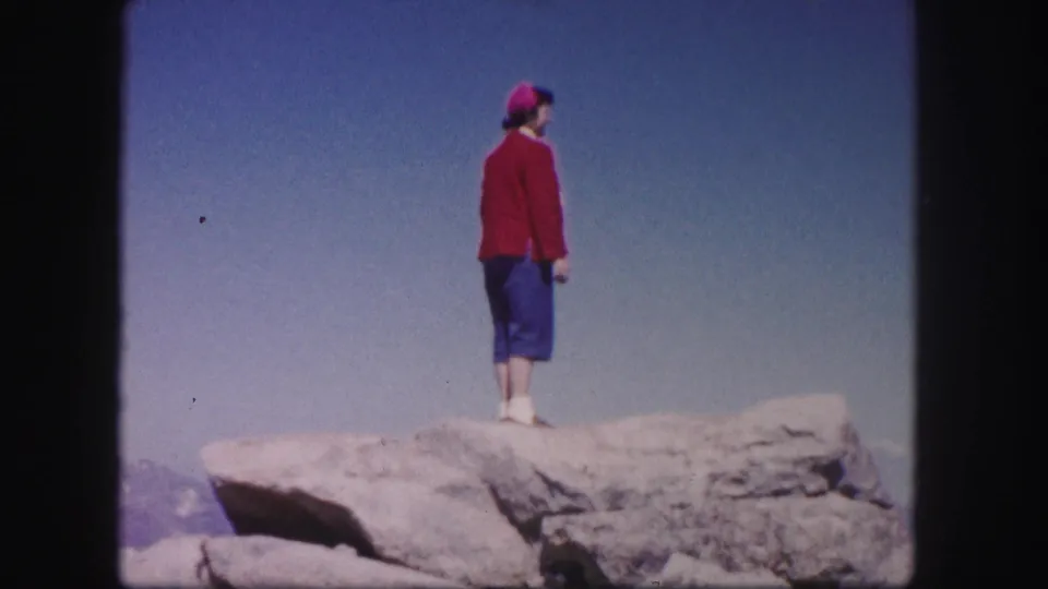 A woman in red and blue stands on a broad rocky summit area in Colorado, filmed on 8mm in 1958.