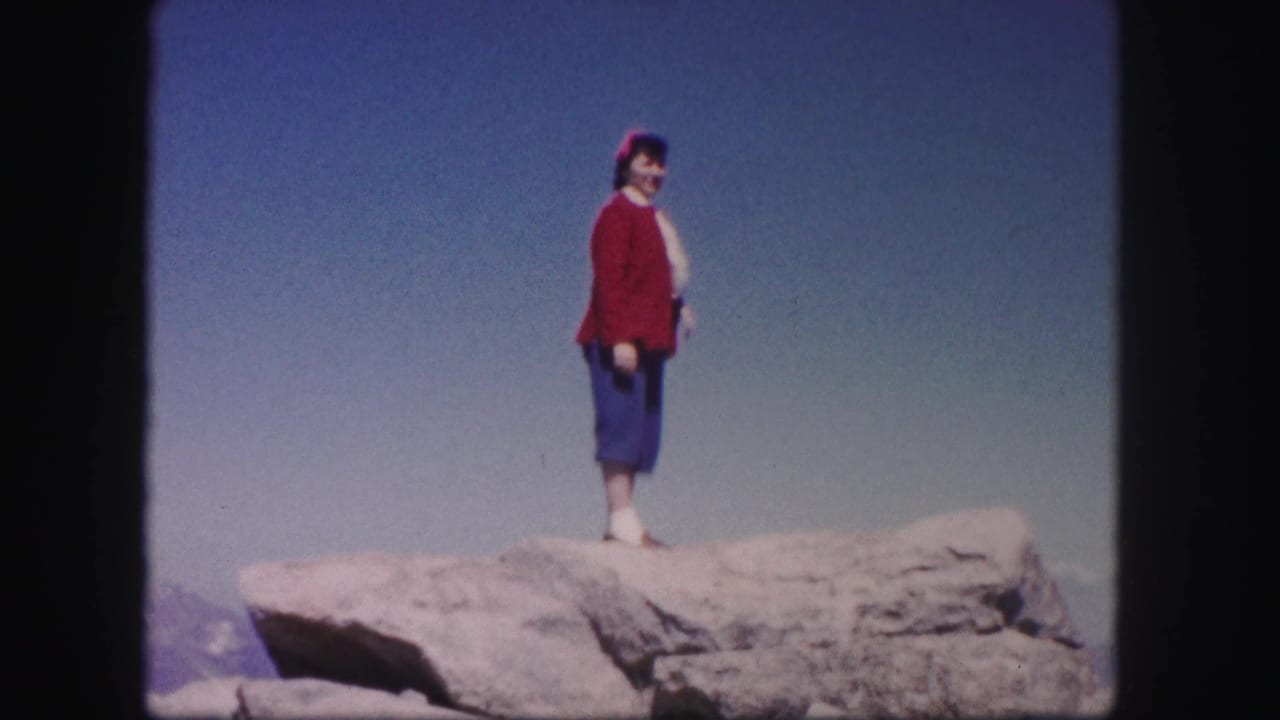 Woman in a red sweater stands on a rocky outcrop under a blue sky.