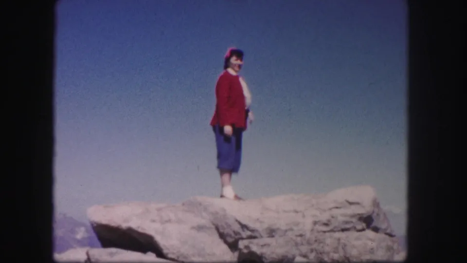 In 1958 Colorado home-movie footage, a woman in a red sweater poses on a pale rocky ledge against open sky.