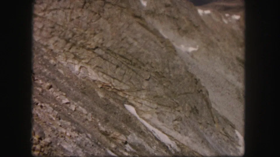 A handheld 1958 Colorado mountain close-up shows rough stone layers, scree, and thin white snow patches on a slope.
