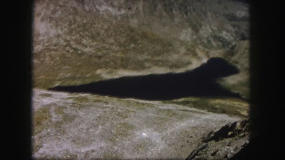 1958 Colorado 8mm frame with a dark elongated small lake or shadowed water shape set among rugged mountain ground.