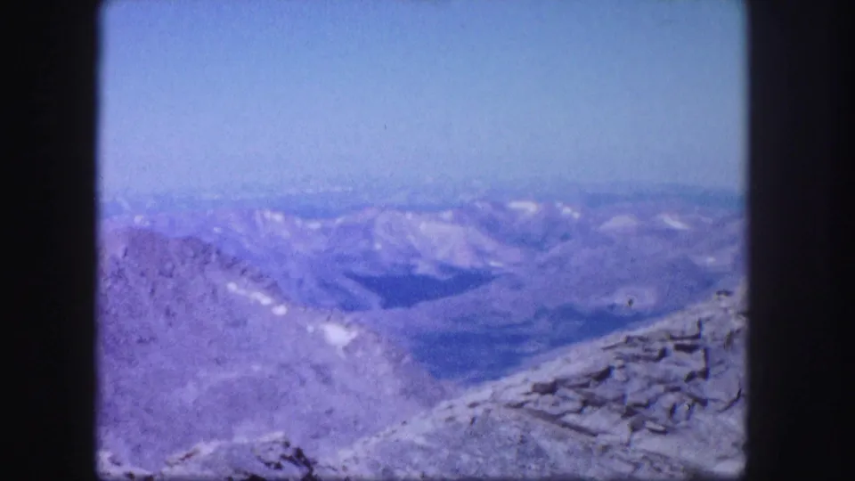 Wide 1958 Colorado mountain panorama with distant ridgelines, snow-dusted areas, rocky foreground, and clear blue sky.