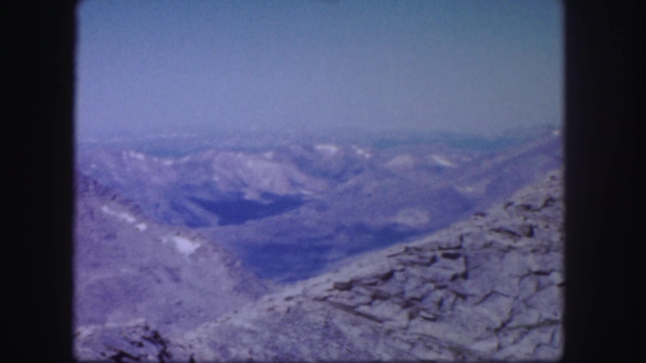 Rocky mountain ridge with distant snow-patched peaks under a pale blue sky.