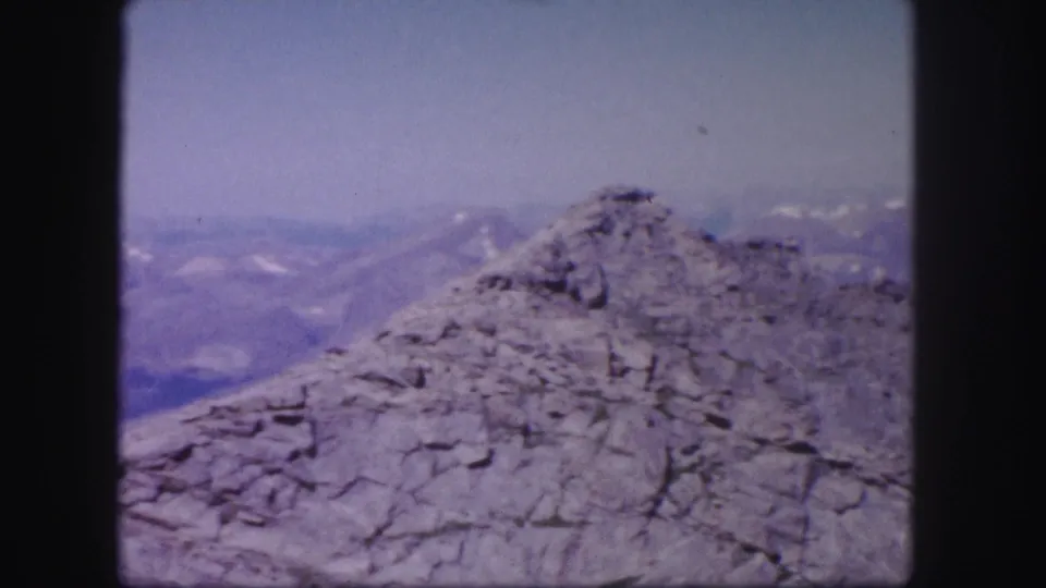 1958 Colorado home-movie frame showing a close rocky summit area against distant hazy mountain terrain.