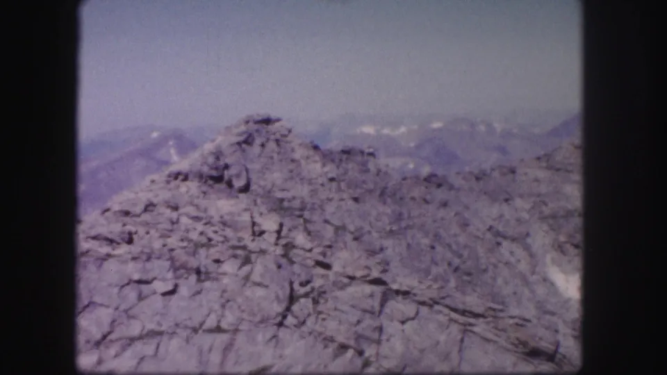 1958 Colorado 8mm frame showing jagged rock in the foreground and hazy mountain ridges with small snow patches in the distance.