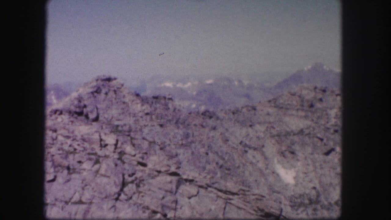1958 Colorado home-movie still with rough foreground rock, bluish distant peaks, and visible 8mm frame edges.