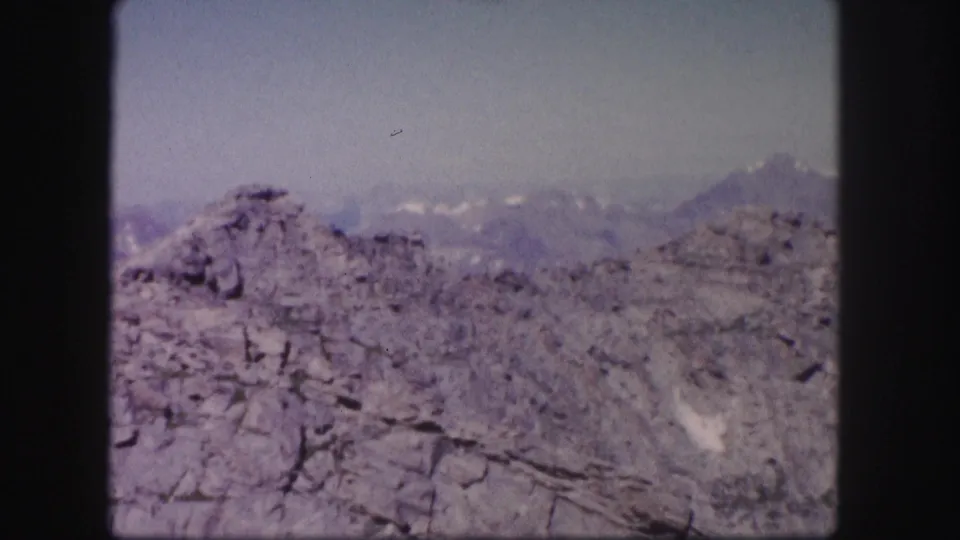 1958 Colorado home-movie still with rough foreground rock, bluish distant peaks, and visible 8mm frame edges.