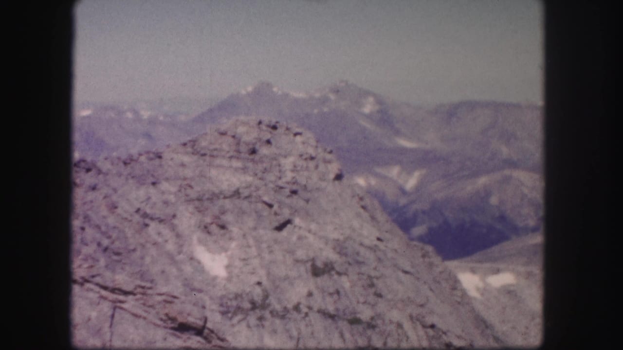 1958 Colorado 8mm frame showing a rocky summit edge, distant mountain peaks, snow patches, and pale daylight sky.