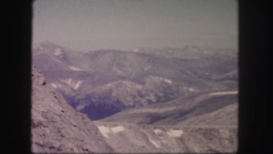 Colorado 1958 8mm frame showing a broad mountain valley, distant ridges, scattered snow, and a rocky slope at left.