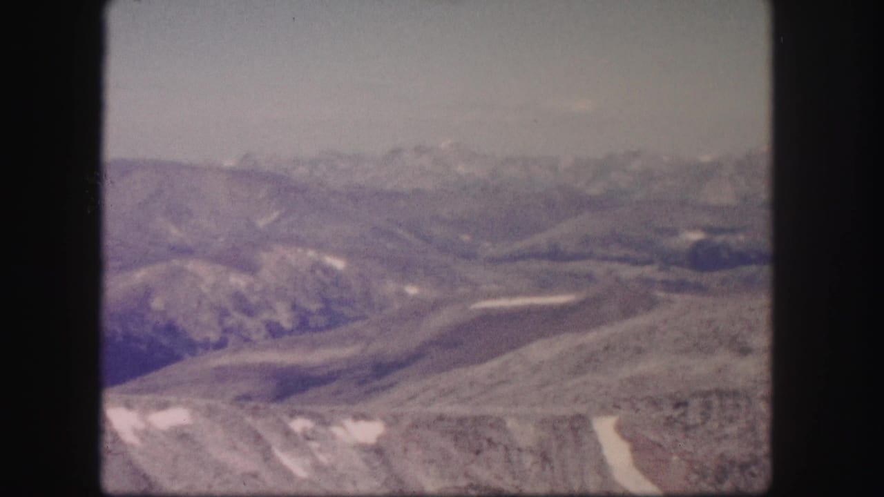 1958 Colorado 8mm frame showing distant hazy mountain ridges, pale sky, and rocky high-country foreground with snow patches.