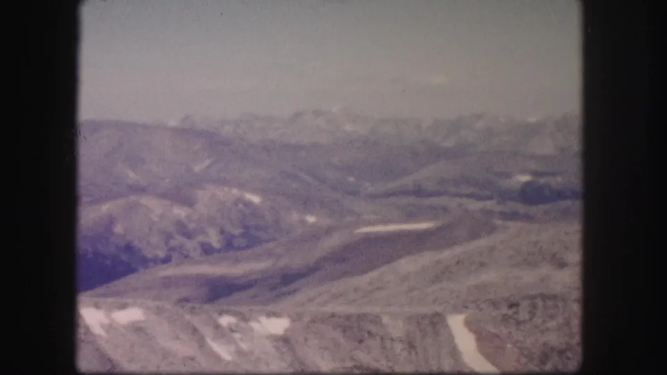 1958 Colorado 8mm frame showing distant hazy mountain ridges, pale sky, and rocky high-country foreground with snow patches.