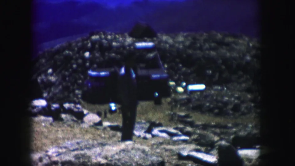 1958 Colorado home-movie frame with a dark-clothed figure standing in rough terrain under a blue-purple cast.