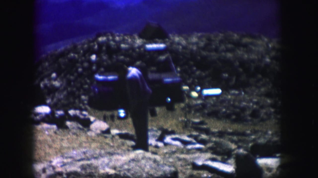 The adult figure appears beside a dark car amid rocks and sparse terrain, with strong blue sky color from the 8mm scan.