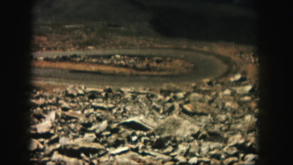 Home-movie frame from 1958 Colorado showing a road bend beyond rocky ground and scrubby terrain.