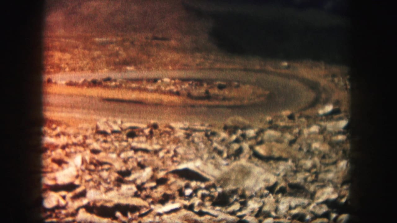 1958 Colorado travel frame with a looping rural road, rocky foreground, dry earth, and warm faded 8mm color.