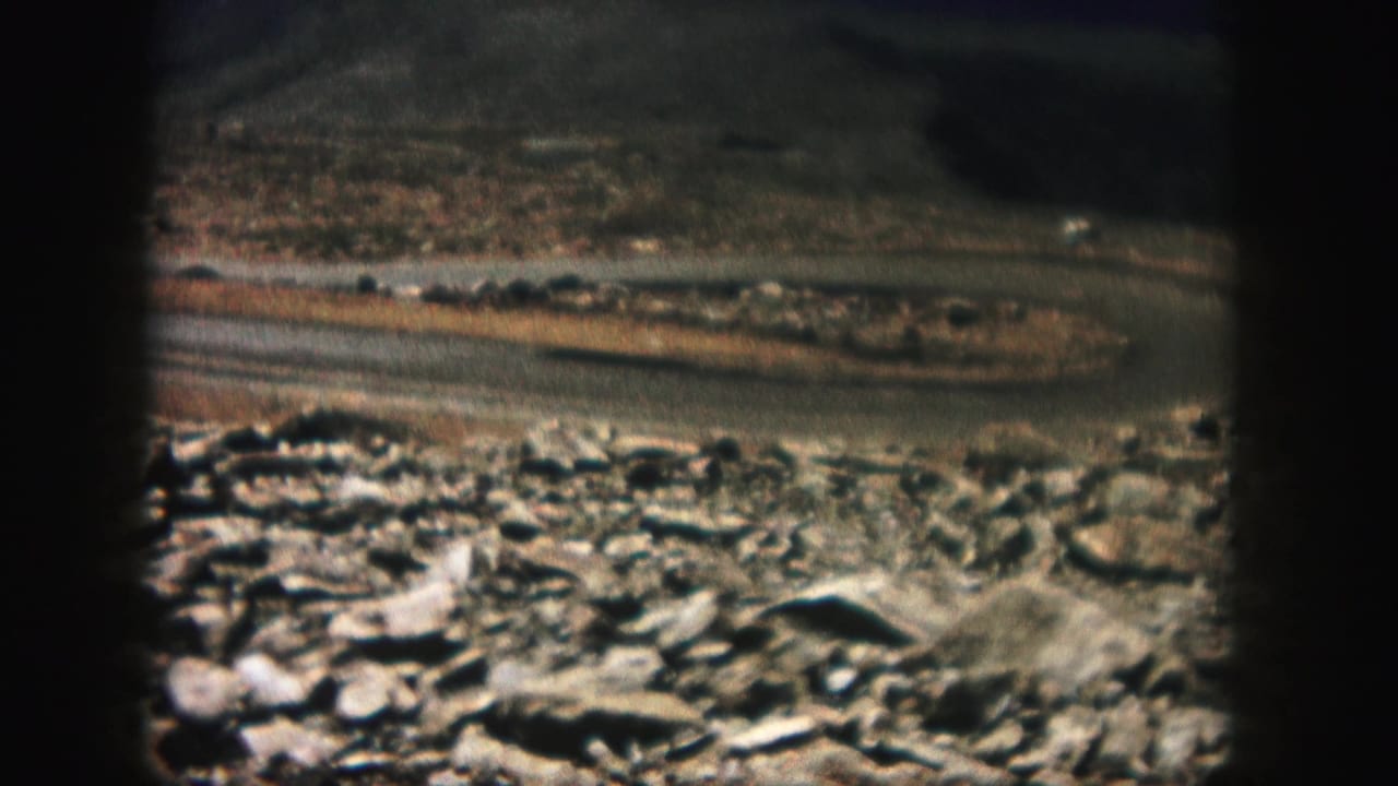 1958 Colorado 8mm frame showing loose rocks in the foreground and a narrow road crossing rugged mountain terrain.